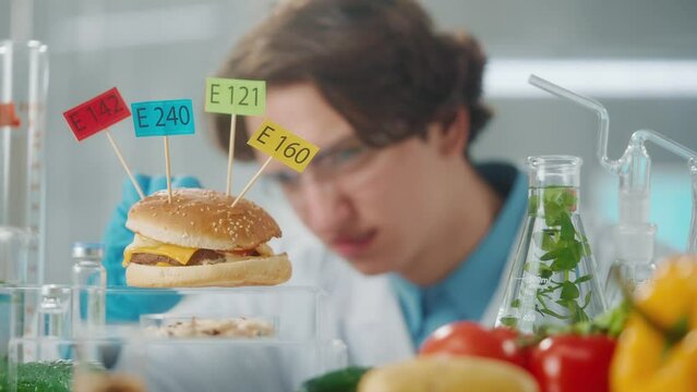 Young male researcher examines a sample of burger labeled E142, E240, E121 and E160. Biolog analyzes the quality of the sample and dissatisfied with the result. Close up.