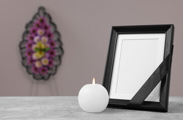 Photo frame with black ribbon, burning candle on table and wreath of plastic flowers near grey...