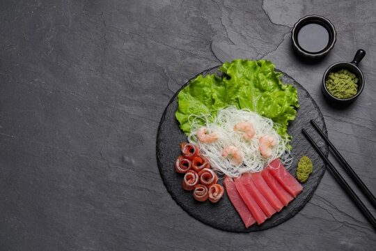 Sashimi set (raw tuna, salmon slices and shrimps) served with funchosa, lettuce, vasabi and soy sauce on dark table, flat lay. Space for text
