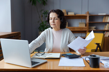 Middle aged senior woman sit with laptop and paper document. Pensive older mature lady reading paper bill pay online at home managing bank finances calculating taxes planning loan debt pension payment