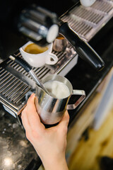 Barista froths milk for coffee. There is a cup of freshly brewed coffee in the coffee machine