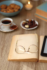 Books, reading glasses, e-reader, plate of chocolate pralines, bowl of cookies, cups of tea and lit candles on the table. Selective focus.