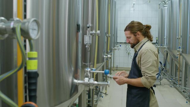 Medium shot of rows of beer fermenting tanks at craft brewery, bearded Caucasian male technician in apron walking in, tapping on pressure gauge, making notes on tablet, and moving on to next one