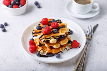 Viennese or Belgian waffles with fresh berries (raspberries and blueberries) and chocolate sauce on a white plate and a cup of coffee. Traditional dessert. Close-up, white background.