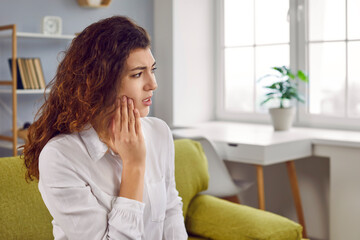 Fototapeta na wymiar Woman suffering from strong teeth pain or feeling discomfort after tooth extraction. Young lady sitting on sofa at home and holding hand on her jaw. Toothache, dental problems concept