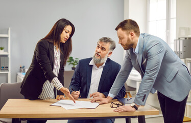 Group of three coworkers and employees at the desk discussing something, reaching agreement and going signing a contract. Confident man with beard on the workplace at office signing some documents.