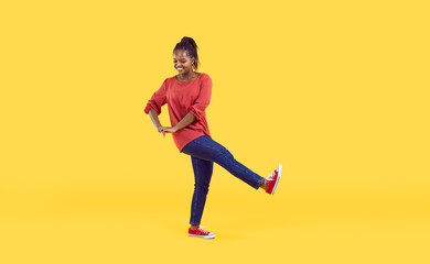 Fototapeta na wymiar Cheerful young afro-american woman having fun isolated on bright yellow background. Smiling woman in casual clothes makes step and various movements looking down. Banner. Full length.