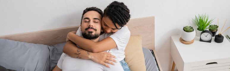 high angle view of tattooed african american woman hugging bearded man lying with closed eyes on bed at home, banner.