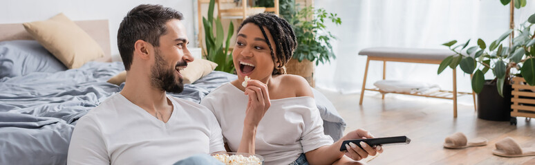 excited bearded man feeding african american woman with popcorn while watching tv in bedroom at home, banner.