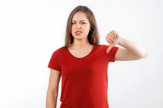 Discontent young woman shows disapproval sign, keeps thumb down, expresses dislike, frowns face in discontent, dressed in red t shirt, standing over white background. Body language concept