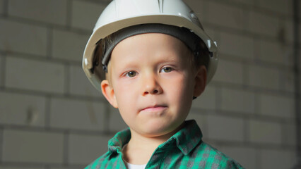 A close-up portrait of a cute little blond boy in a white helmet and a green shirt at a construction site. High quality photo