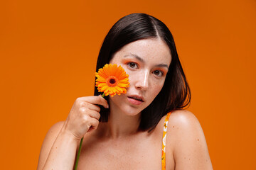 Portrait of young , natural, freckled woman with flower on her face, isolated over orange studio...