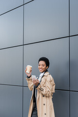 pleased african american businesswoman in trendy trench coat messaging on mobile phone while standing near grey wall on street.