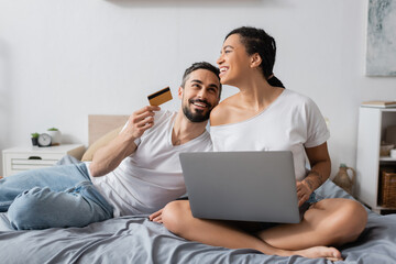 happy bearded man holding credit card near laughing african american girlfriend sitting with laptop on bed at home.