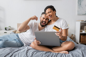 overjoyed interracial couple laughing with closed eyes near laptop and credit card on bed at home.