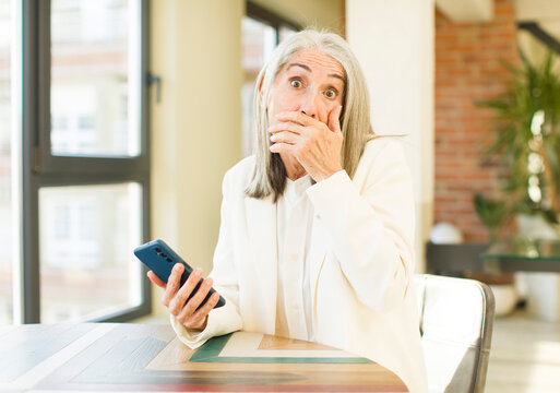 pretty senior woman covering mouth with a hand and shocked or surprised expression with a smartphone