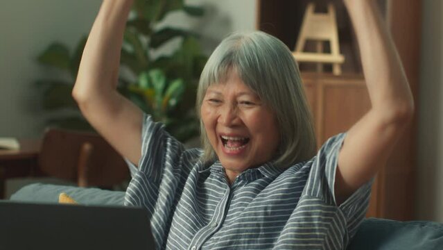 Happy attractive Asian elderly woman smile checking email received good news, feeling surprised. Cheerful old beautiful aged female looking at screen sit on sofa  in home living room feeling excited.
