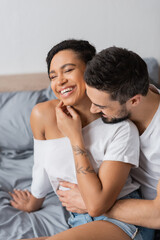 bearded man hugging carefree african american woman smiling with closed eyes in bedroom at home.