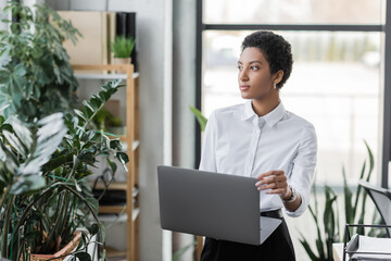 pensive african american businesswoman with laptop looking away while standing near green plants in modern office.