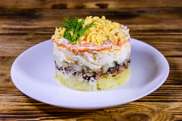 Plate with traditional russian salad Mimosa on a wooden table. Layered salad with potatoes, sardine, cheese, carrot, eggs and mayonnaise