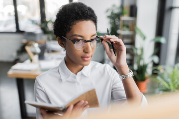 african american businesswoman in white blouse adjusting eyeglasses while holding notepad and pen in office.