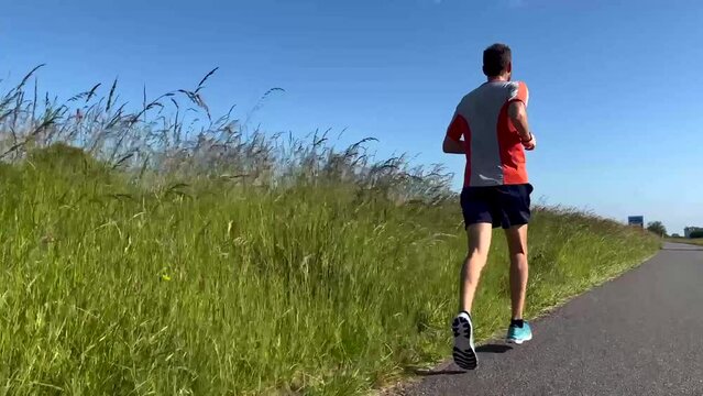 Low angle closeup of legs following a male trail runner on floodplains valley dyke asphalt road. Athlete endurance training run exercise outdoor sports concept.