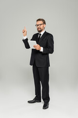 full length of businessman in formal wear and glasses holding digital tablet while pointing with finger on grey.