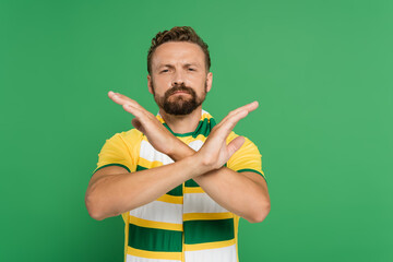 bearded soccer fan in striped scarf and yellow t-shirt showing stop gesture with hands isolated on green.