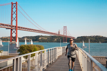 Man running across the bridge in front of a beautiful city landscape. Lisbon promenade, The 25 de Abril Bridge, The Sanctuary of Christ the King, sailboats. Healthy lifestyle