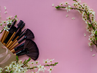 Bouquet of cosmetic brushes in paper shopping bag on pink background with cherry flowers and copy space. Women and Mothers Day gift. Concept of beauty and femininity. Womens secrets. Tools for make up