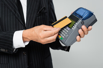 cropped view of businessman in suit holding credit card near payment terminal isolated on grey.