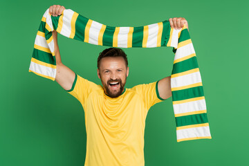excited soccer fan in yellow t-shirt holding striped scarf and looking at camera isolated on green.