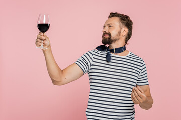 cheerful man in striped t-shirt holding glass of red french wine isolated on pink.