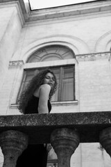 A beautiful portrait of a curly brunette girl in a black dress and makeup, poses gorgeous on balcony, looking at camera. Black and white photo.