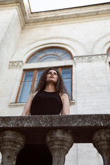 A beautiful portrait of a curly brunette girl in a black dress and makeup, poses gorgeous on balcony, looking up. Beauty portrait. 