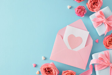 Top view photo of envelope postcard with heart natural flowers pink rose buds gift boxes and small hearts baubles on light blue background with empty space. Mother Day concept