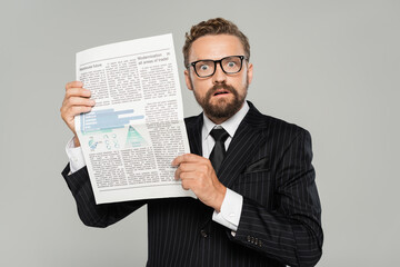 shocked businessman in suit and glasses holding newspaper isolated on grey.