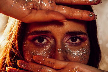 Close-up brown eyes of a young beautiful woman, makeup with sparkles, hands on face.