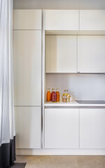 
vertical frame with a view of the interior of the kitchen, kitchen white facades, a curtain, an induction stove, bottles with liquid, juice or pickles