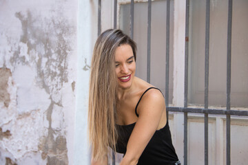 Fototapeta premium Young blonde woman dressed in black t-shirt and jeans leaning against an old wall. The woman is happy and smiling. Yellow day concept.