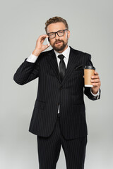 Naklejka premium businessman in glasses and suit talking on smartphone and holding paper cup isolated on grey.