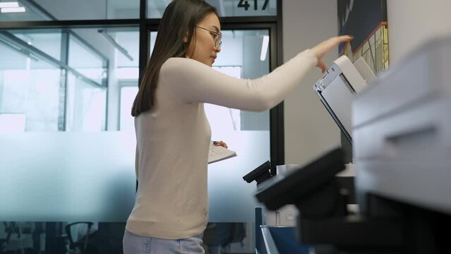 Handsome asian woman wearing eyeglasses prints documents on printer in office