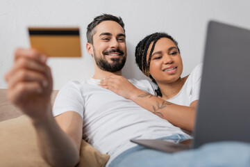 interracial couple with laptop and credit card smiling during online shopping on blurred foreground in bedroom at home.