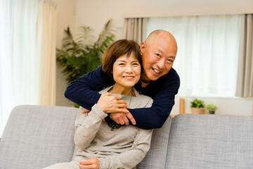 ソファに座る妻に後ろから抱きつく夫（シニアの夫婦の愛情表現）