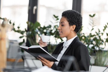 side view of african american businesswoman sitting with notebook and coffee cup in office.