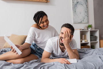 excited man covering face and laughing near african american girlfriend with digital tablet on bed at home.