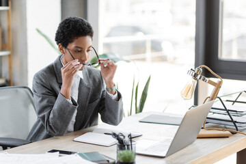 stylish african american businesswoman putting on eyeglasses while sitting near laptop and blank notebook in office.