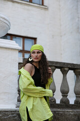 The beautiful brunette with curly hair, wears a green scarf on her head and shirt, poses outside, looking at the camera, sunset background.