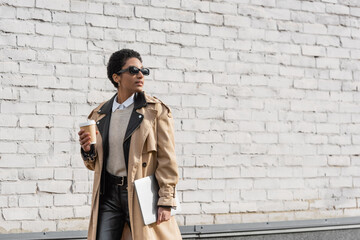 african american businesswoman in sunglasses and beige trench coat walking with laptop and coffee to go along brick wall on urban street.