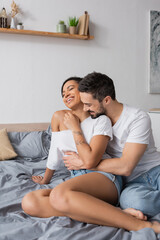 bearded man in white t-shirt hugging carefree african american woman sitting on bed at home.